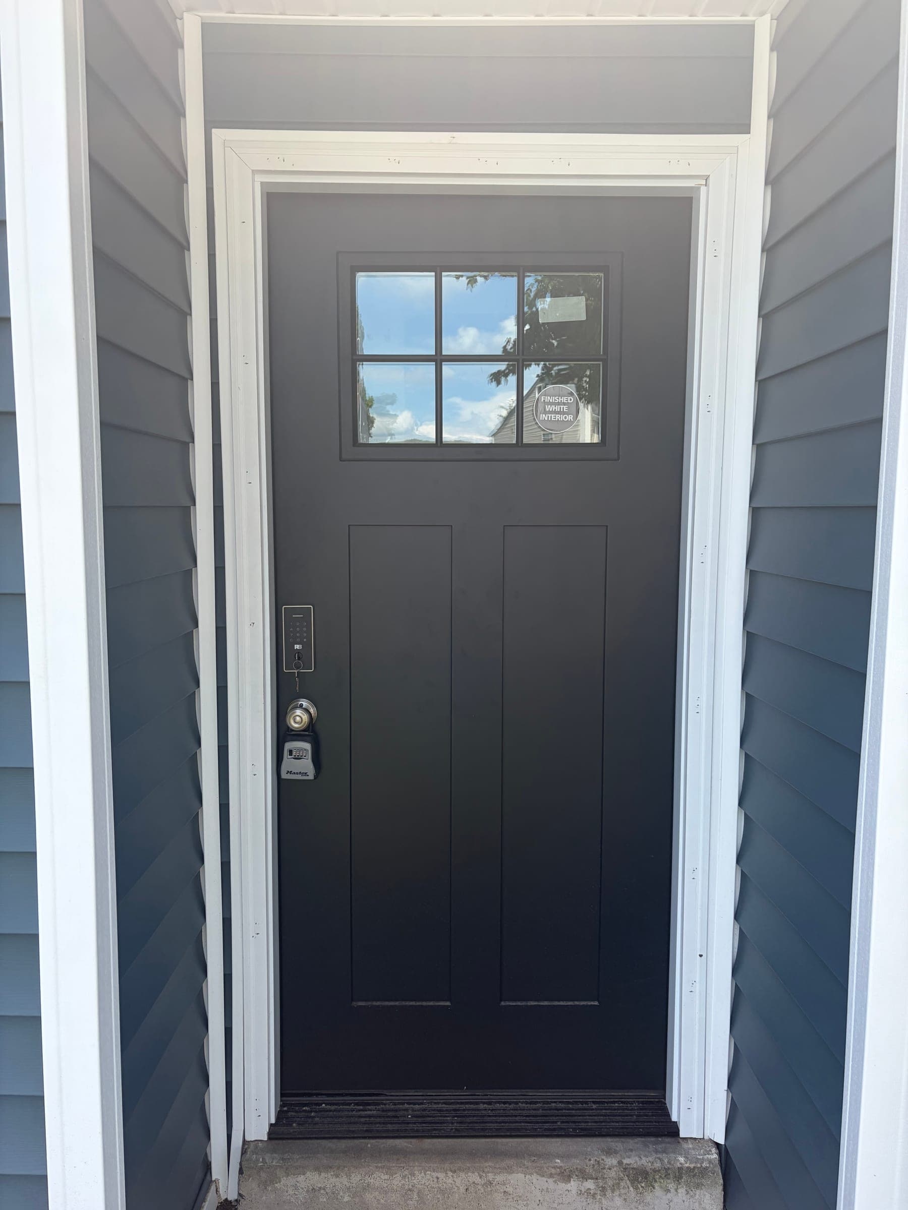 New black craftsman-style entry door with a six-lite window, installed with fresh white trim on a gray-sided home