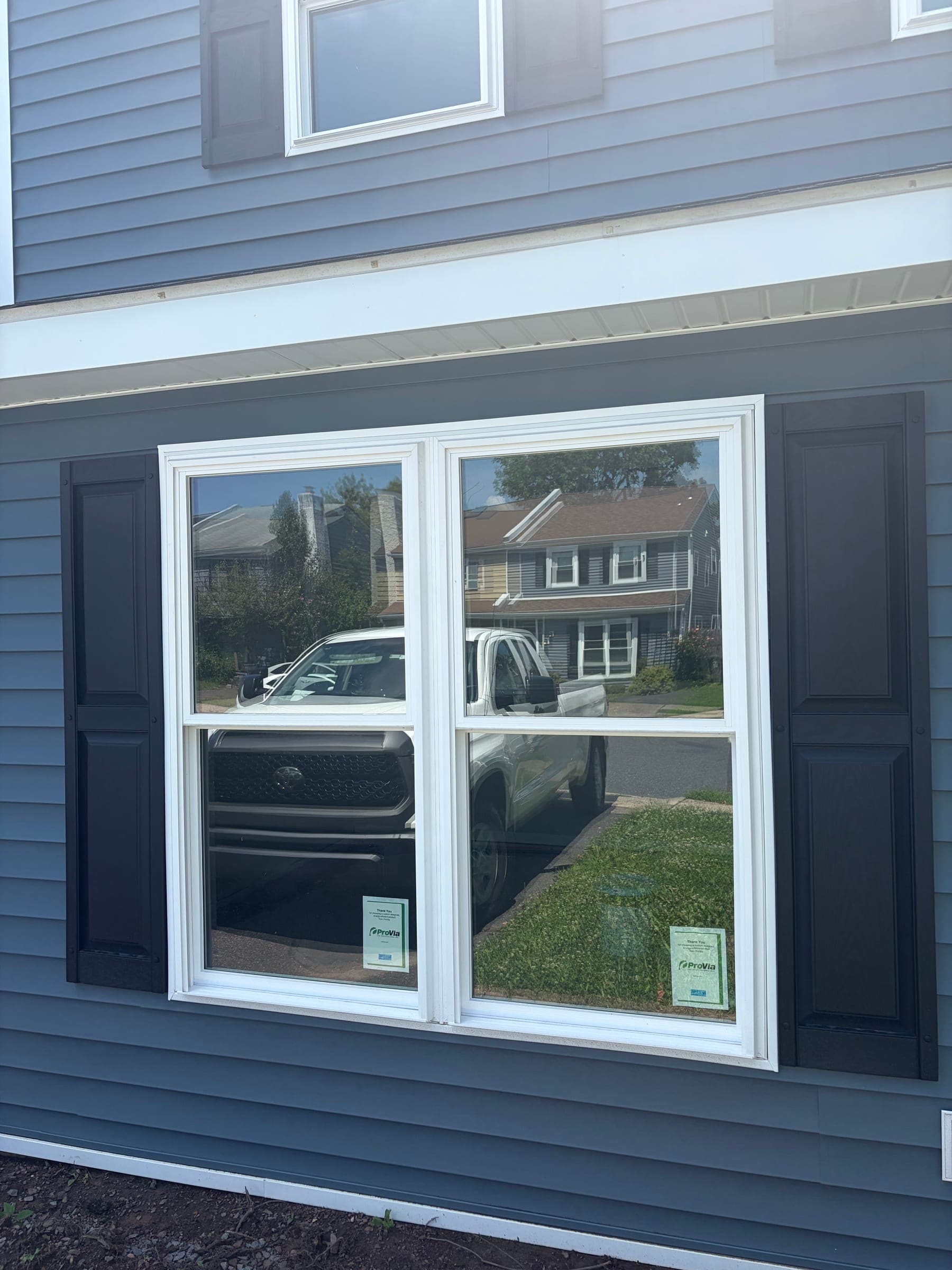 Twin double-hung windows with white trim and black shutters installed on a gray-sided home